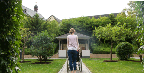 Mitarbeiterin schiebt Bewohner des Seniorenzentrums St. Hedwig im Rollstuhl durch den Garten